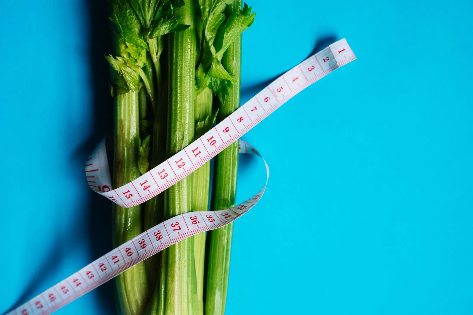 Tape measure wrapped around fresh celery on a blue background, representing healthy weight management