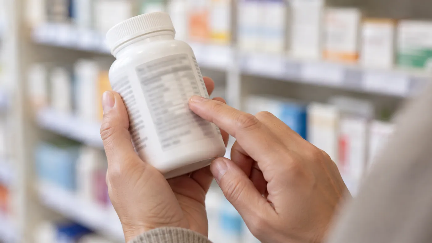 Magnesium Complex vs Single-Form: Which Blend Is Worth It 3 Hands holding a supplement bottle in a pharmacy aisle with a finger on the Supplement Facts panel, checking the magnesium amounts